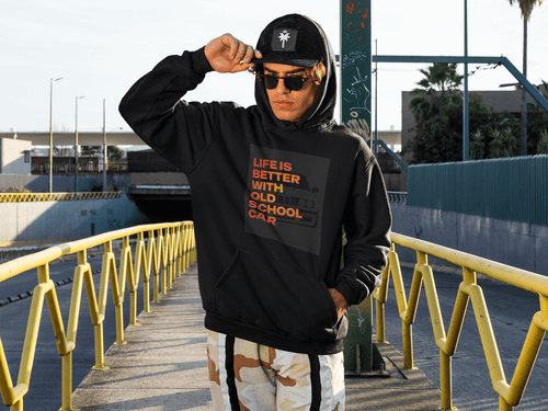 Person wearing a black hoodie with text Life is better with old school car and graphics BMW e30, standing on a bridge with yellow railings. Bavarian old school