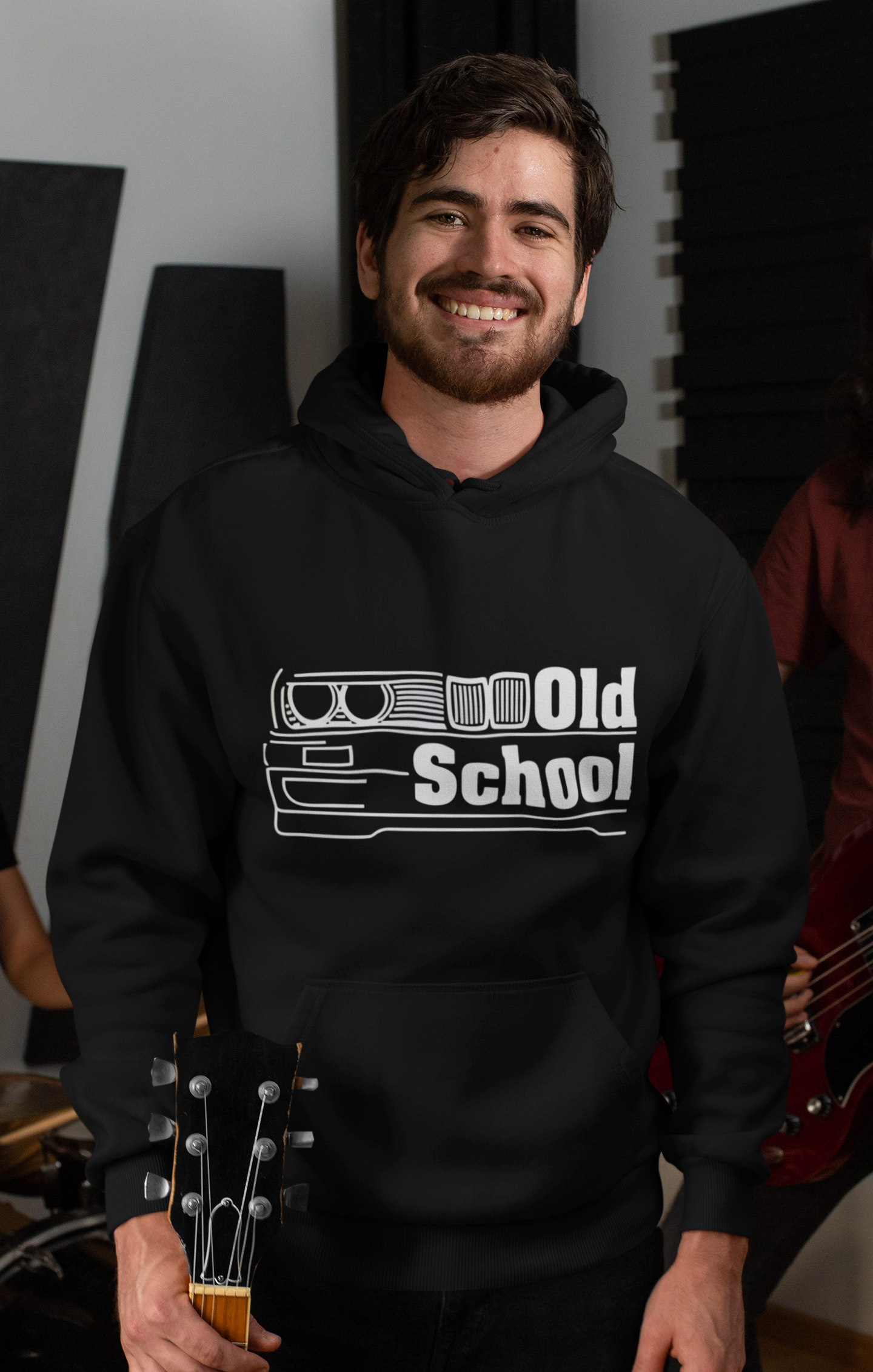 Man wearing a black hoodie with 'Old School' design, holding a guitar in a room with musical equipment.