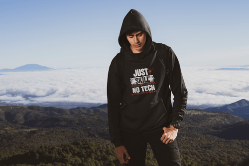 Person wearing a hoodie with text Just Skill no tech, standing on a mountain with a scenic view. Bavarian old school