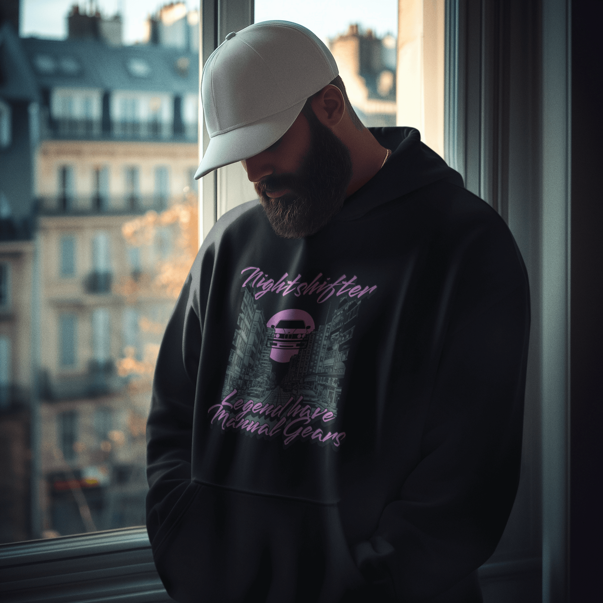 Person wearing a black hoodie with text and graphics, standing by a window with cityscape view.