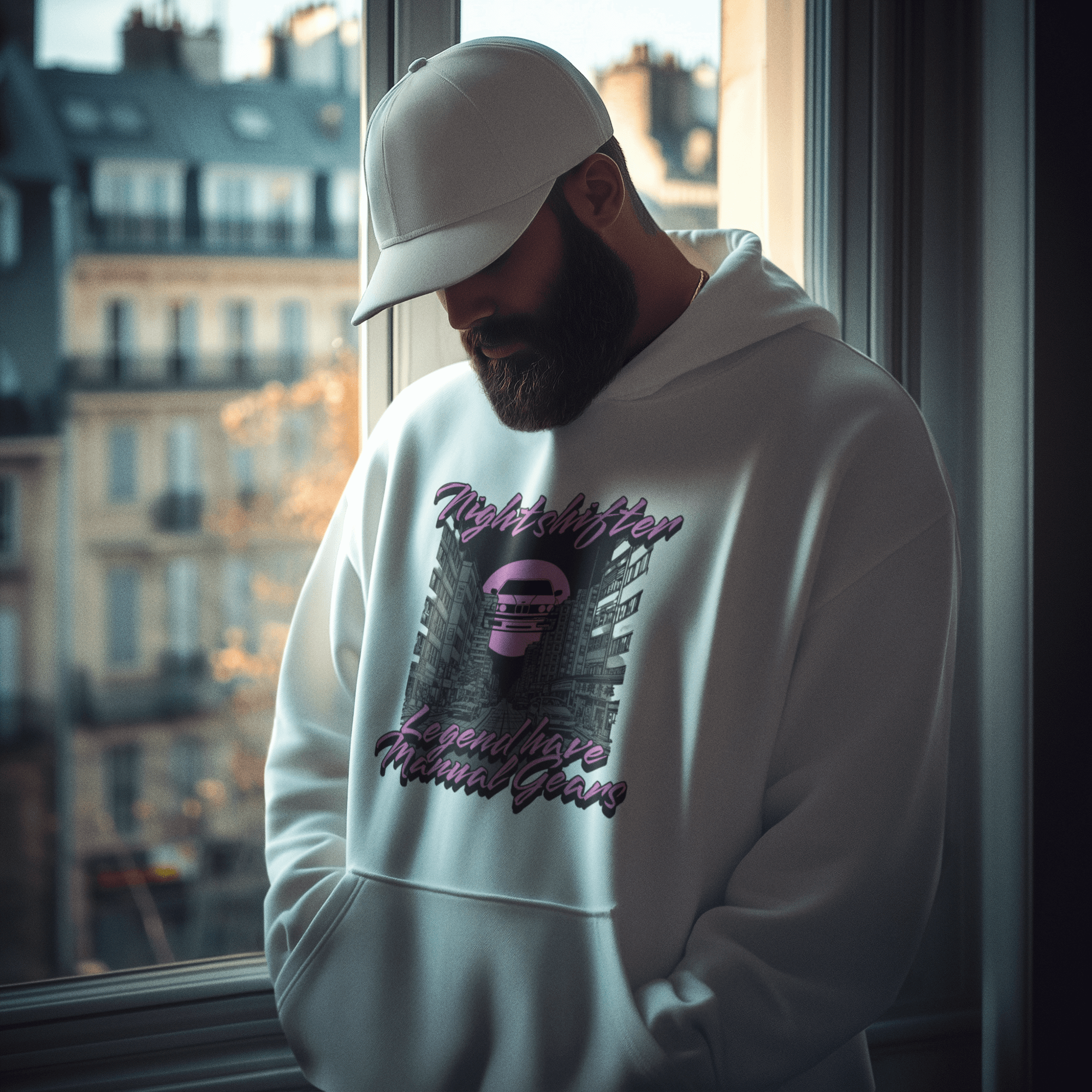 Man wearing a white hoodie with graphic design and text, standing by a window with cityscape view.