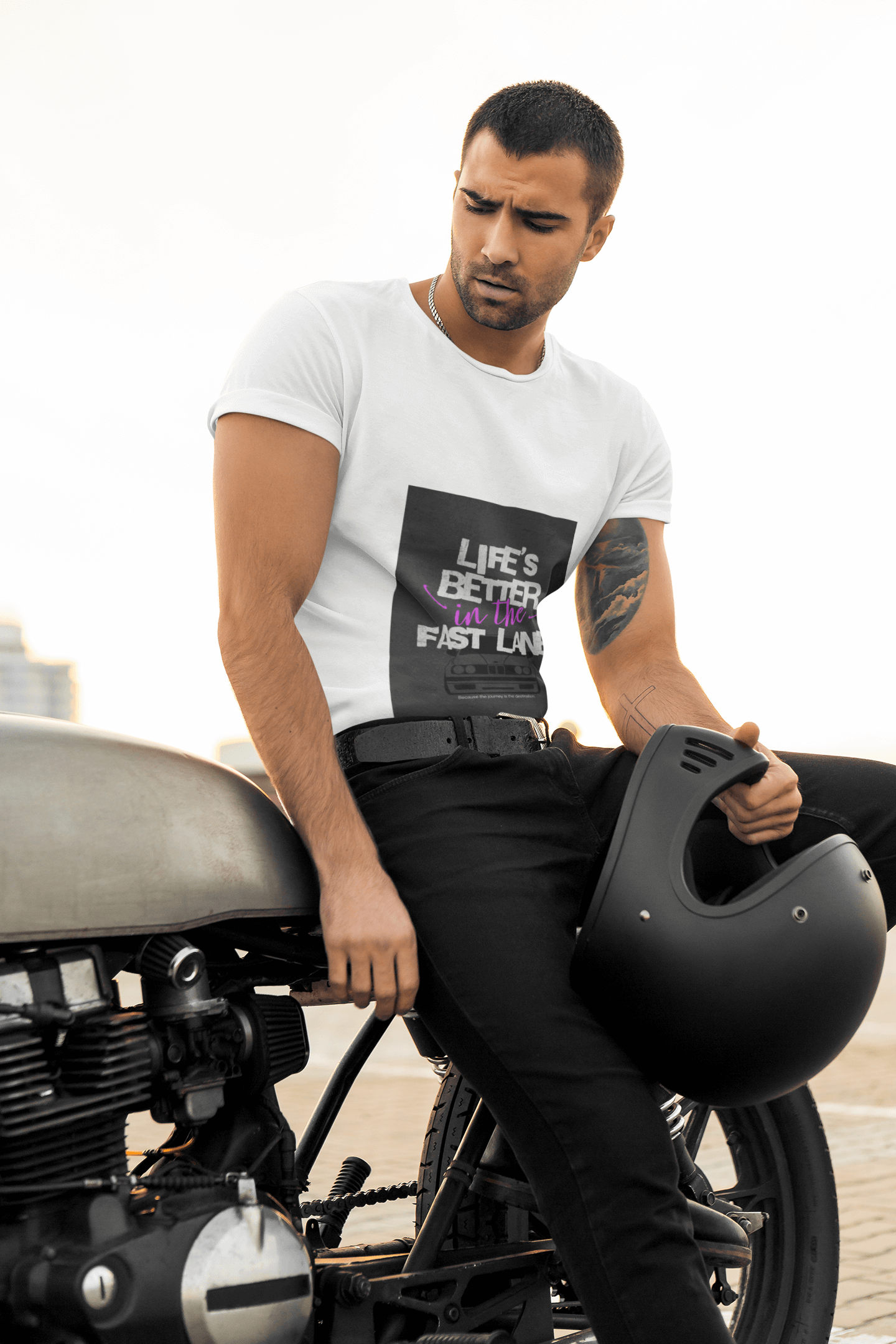 Man sitting on a motorcycle holding a helmet, wearing a white t-shirt with text Life`s better in the fast lane. bavarian old school